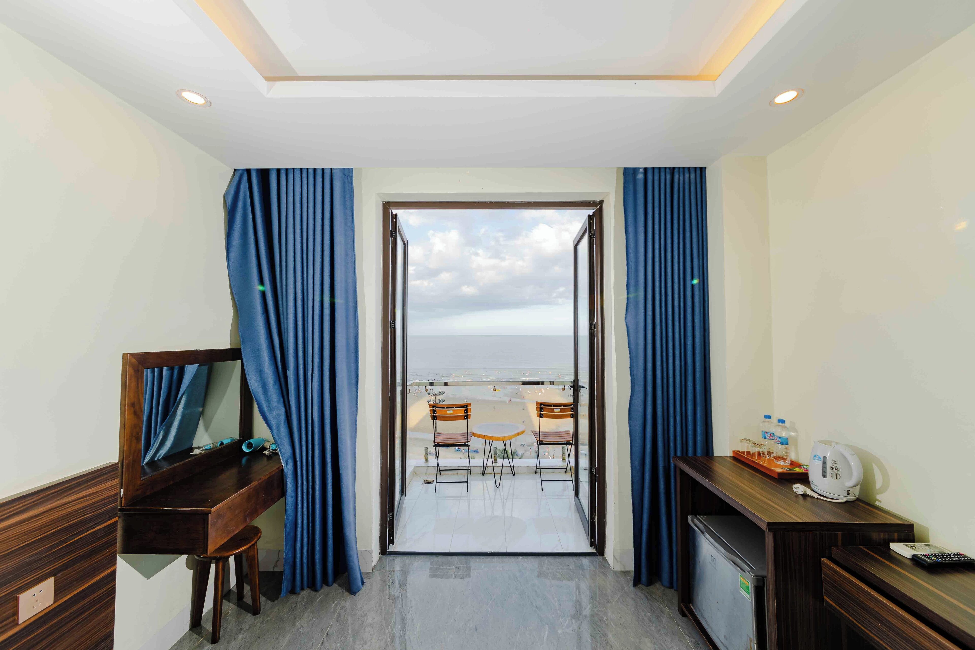 A hotel room with a desk, a chair, a small refrigerator, and a view of the beach through an open door.