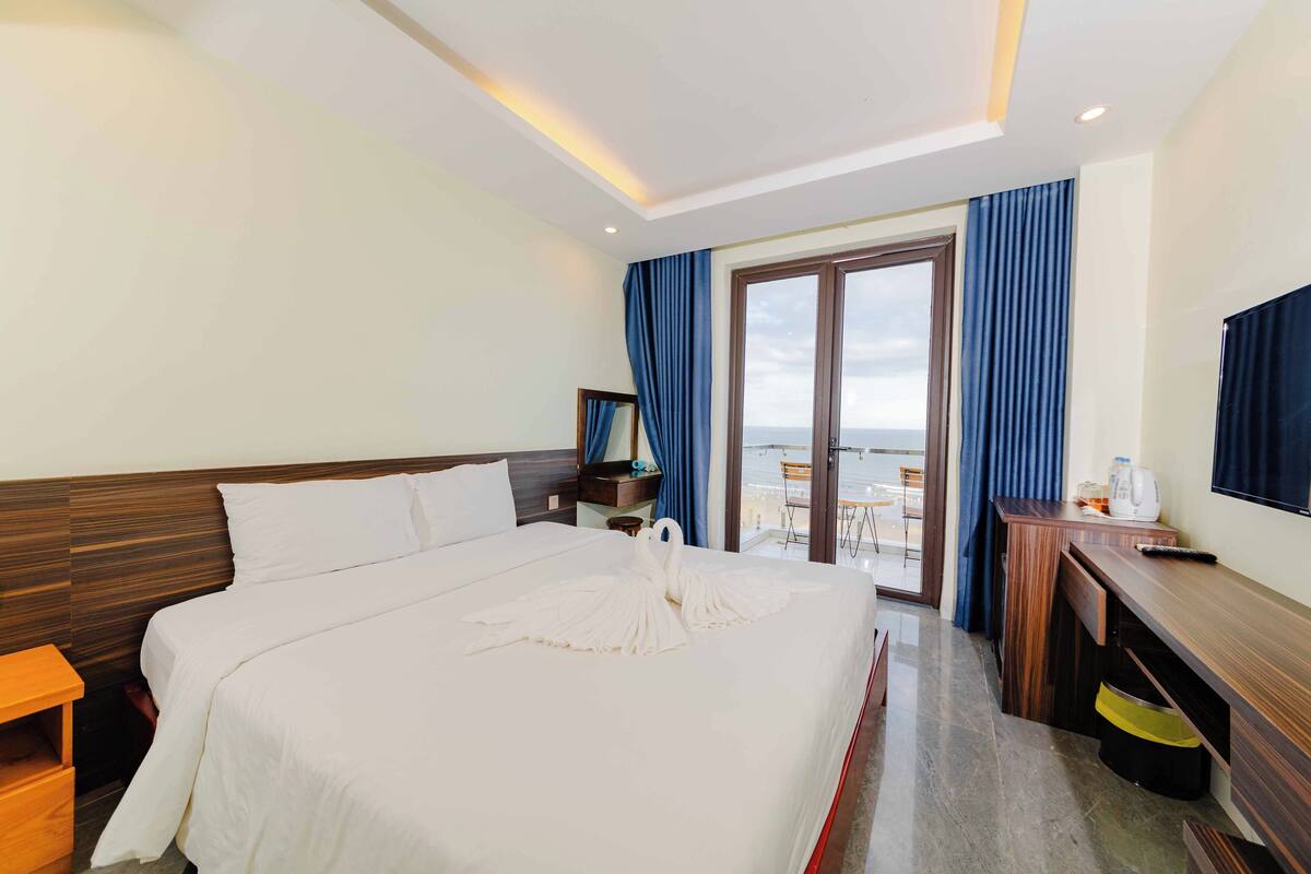 Ein Hotelzimmer mit einem großen Bett, einem Schreibtisch, einem Sessel und Meerblick durch eine Glastür.
