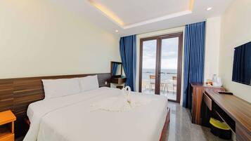 A hotel room with a large bed, a desk, a chair, and a view of the sea through a glass door.