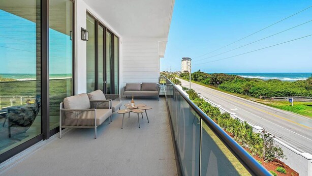 A balcony with a sofa, chairs, and a small table, overlooking a road and greenery with a clear view of the ocean. - Hi3051hl202: 3 Bed, Ocean Breeze (Melbourne Beach)