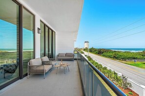 Ein Balkon mit einer Couch, Stühlen und einem kleinen Tisch, mit Blick auf eine Straße und Grünflächen, mit freiem Meerblick.