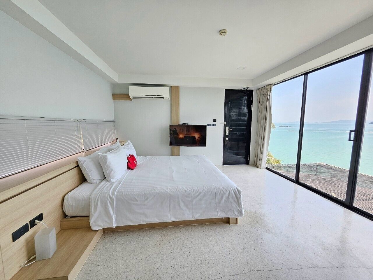 A modern bedroom with a large bed, a flat-screen TV, and a view of the sea through large windows.