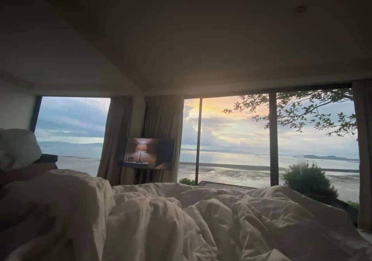 A bedroom with a large bed, a TV on a stand, and a view of the sea through large windows.
