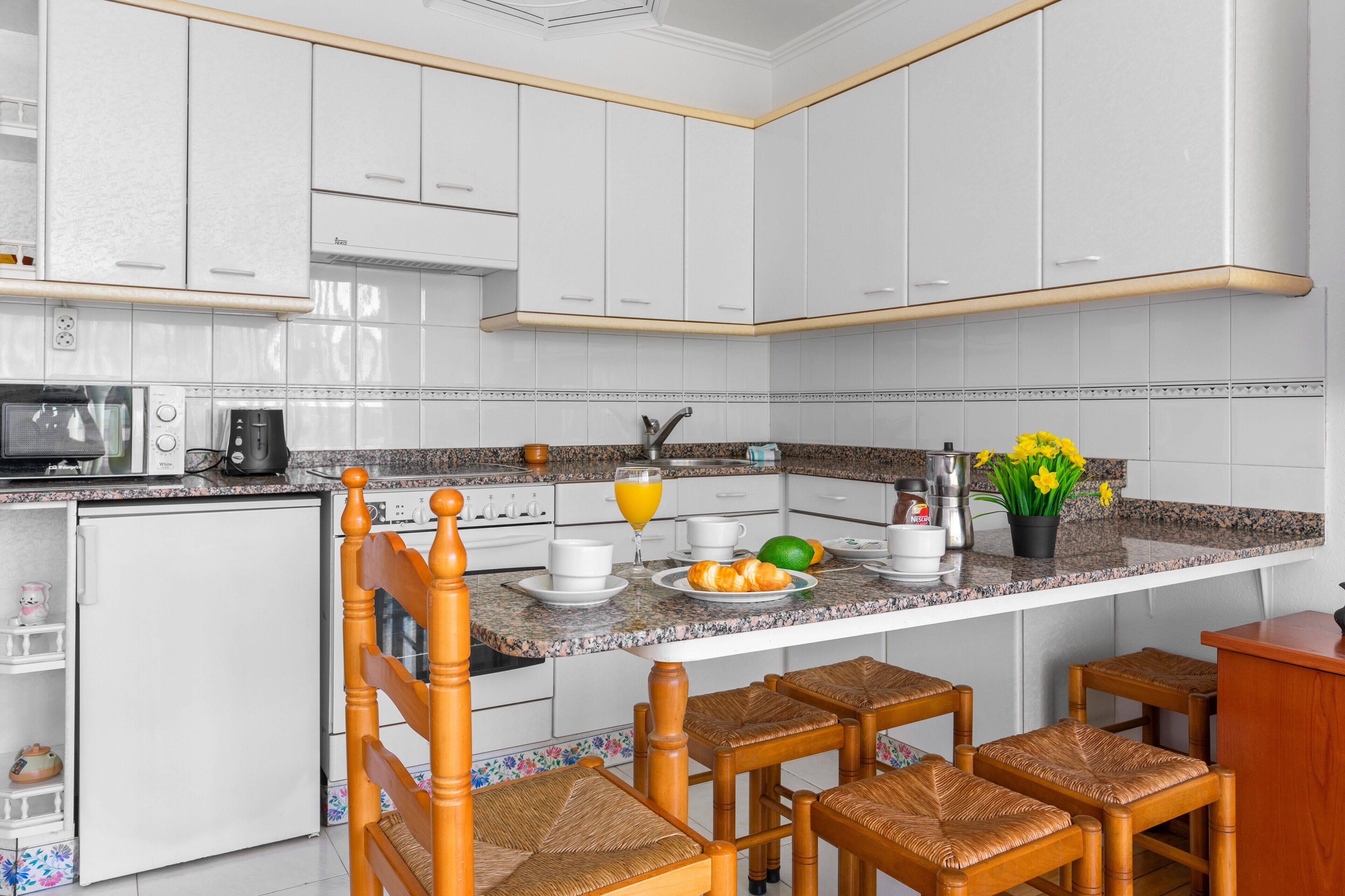 Uma cozinha com armários brancos, bancada de granito, geladeira, micro-ondas, máquina de café e um espaço para café da manhã com croissants, suco de laranja e flores.