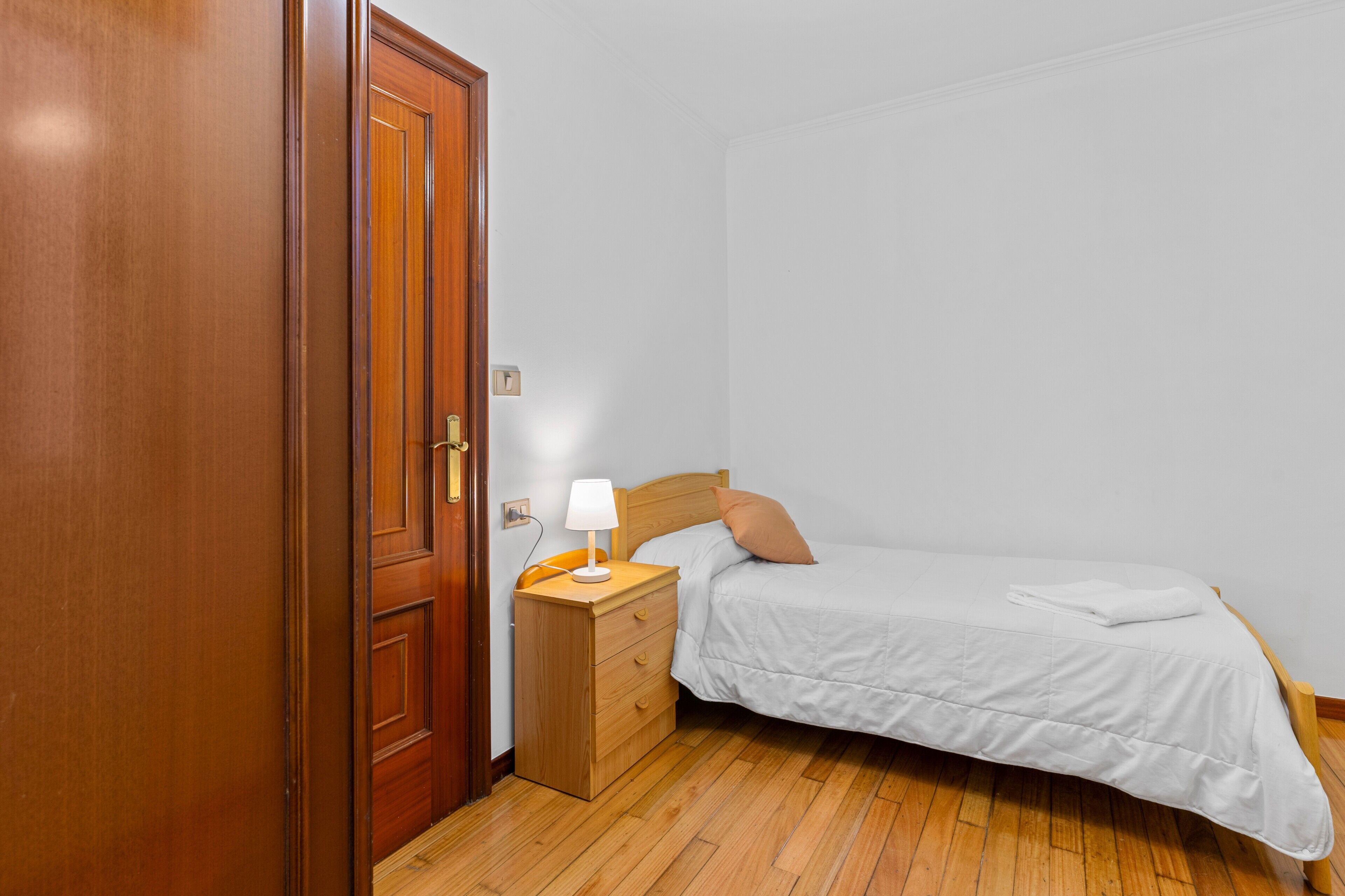 Een slaapkamer met een houten deur, een nachtkastje met een lamp, een bed met een witte dekbedovertrek en een houten vloer.