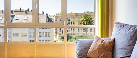 Una habitación con un ventanal grande, un sofá con un libro y vistas a edificios y una palmera.