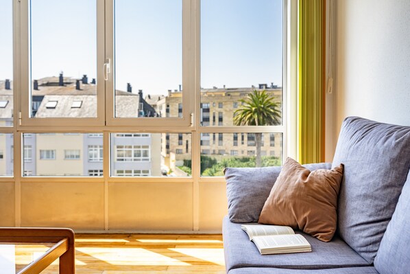 Une pièce avec une grande fenêtre, un canapé sur lequel repose un livre, et une vue sur des immeubles et un palmier.