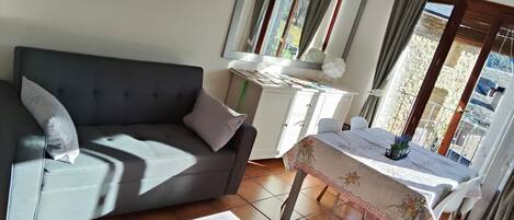 Una habitación con un sofá gris, una mesita blanca, un armario blanco y una ventana con cortinas.