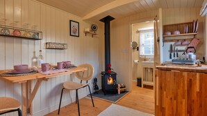 Une cuisine chaleureuse aux murs en bois, avec un poêle à bois central, un coin repas comprenant une table et des chaises, et des étagères remplies d’ustensiles de cuisine.
