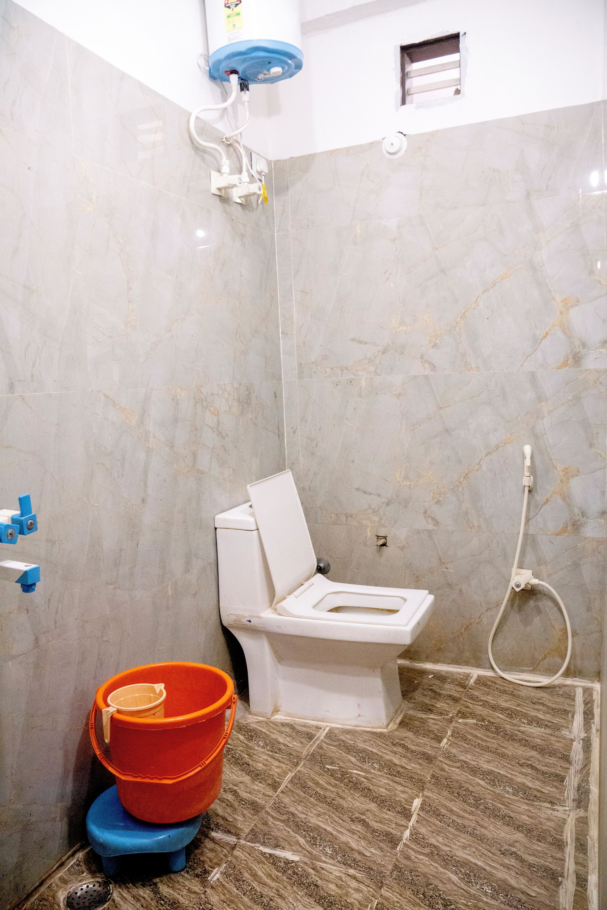 A bathroom with a toilet, shower, and a small window. There are two buckets, one orange and one blue, placed on the floor.