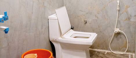 A bathroom with a toilet, shower, and a small window. There are two buckets, one orange and one blue, placed on the floor.