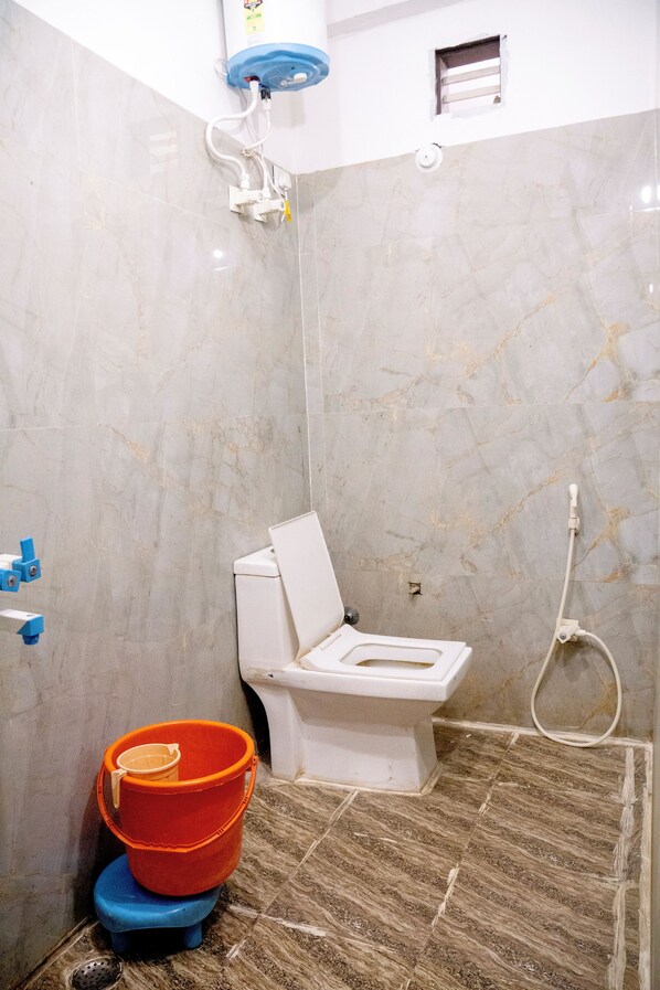 Ein Badezimmer mit Toilette, Dusche und einem kleinen Fenster. Zwei Eimer, ein orange und ein blauer, stehen auf dem Boden.