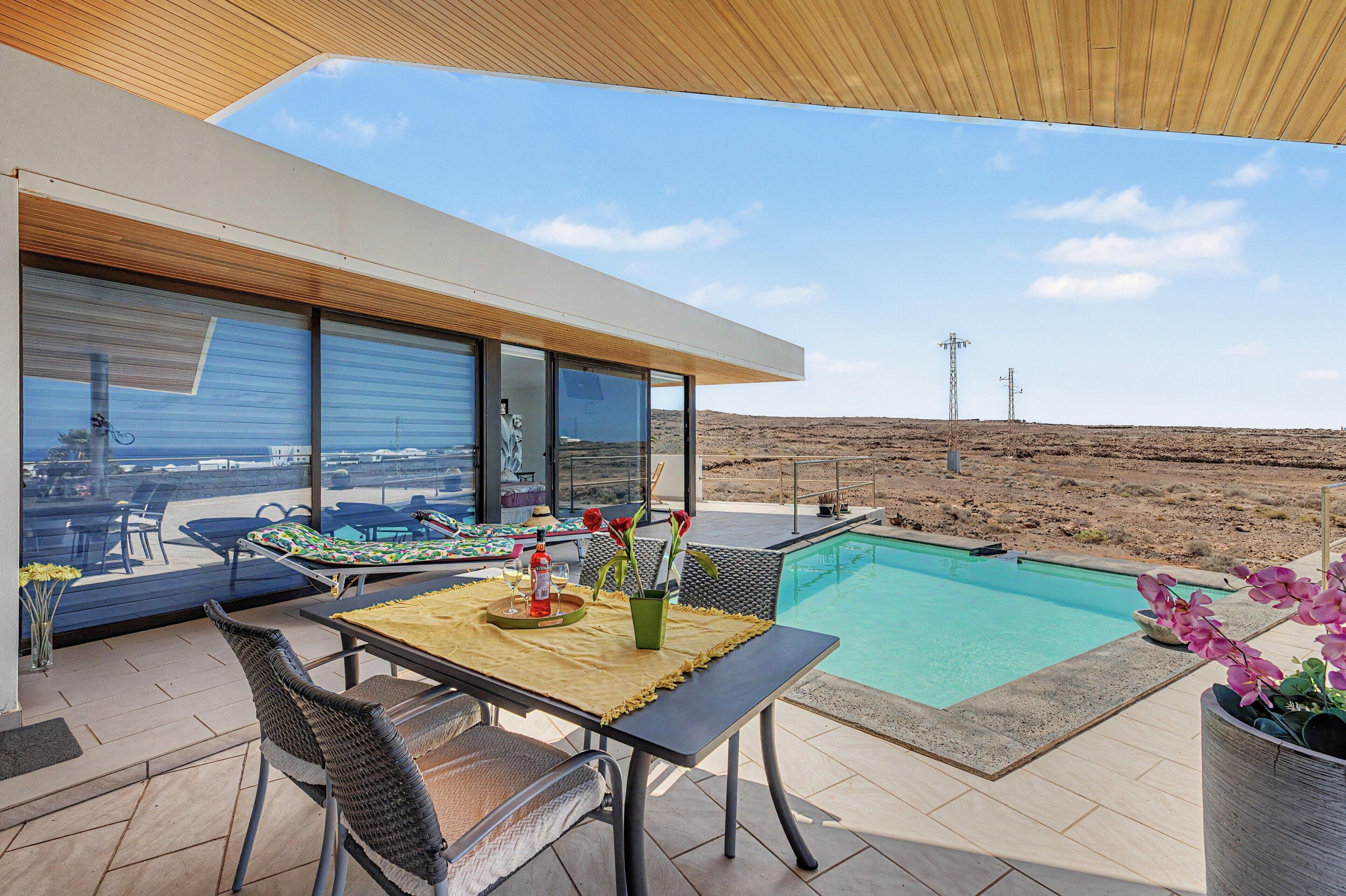 Un área de piscina moderna al aire libre con una mesa de comedor y sillas, una silla de descanso y vistas al paisaje desértico.