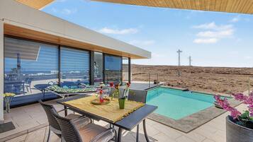 Un área de piscina moderna al aire libre con una mesa de comedor y sillas, una silla de descanso y vistas al paisaje desértico.