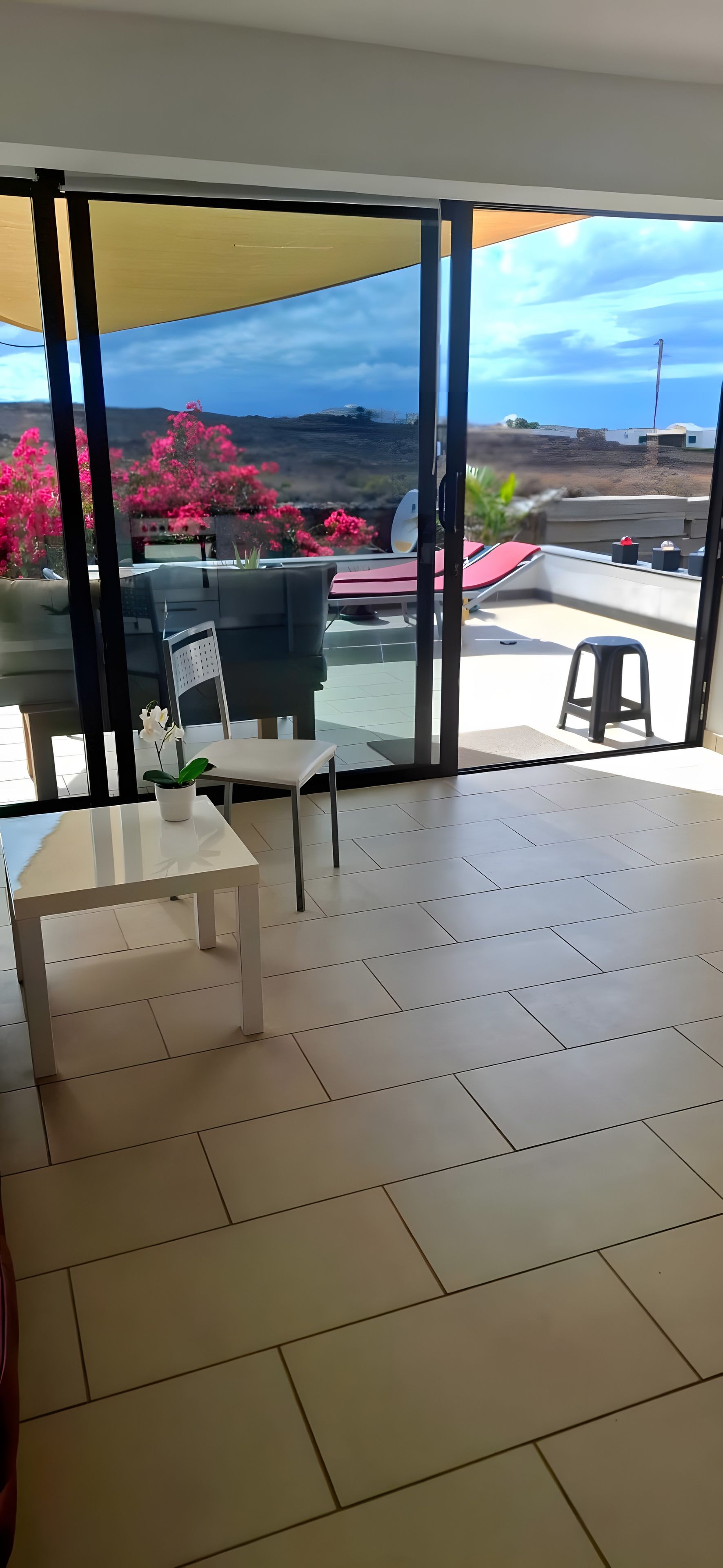 Une pièce dotée d’une grande porte-fenêtre coulissante donnant sur une terrasse extérieure. La pièce est carrelée et comprend une petite table blanche sur laquelle est posé un vase avec une seule fleur.