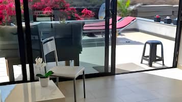 Une pièce dotée d’une grande porte-fenêtre coulissante donnant sur une terrasse extérieure. La pièce est carrelée et comprend une petite table blanche sur laquelle est posé un vase avec une seule fleur.