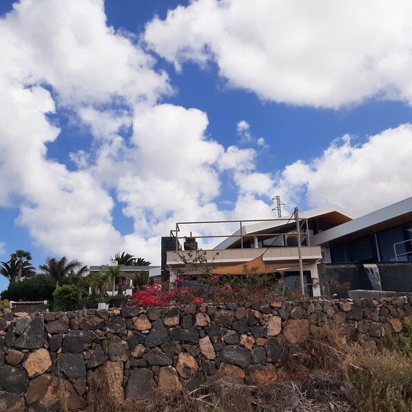 Un bâtiment moderne avec un mur de pierre au premier plan, un ciel bleu dégagé parsemé de nuages et une ligne électrique.