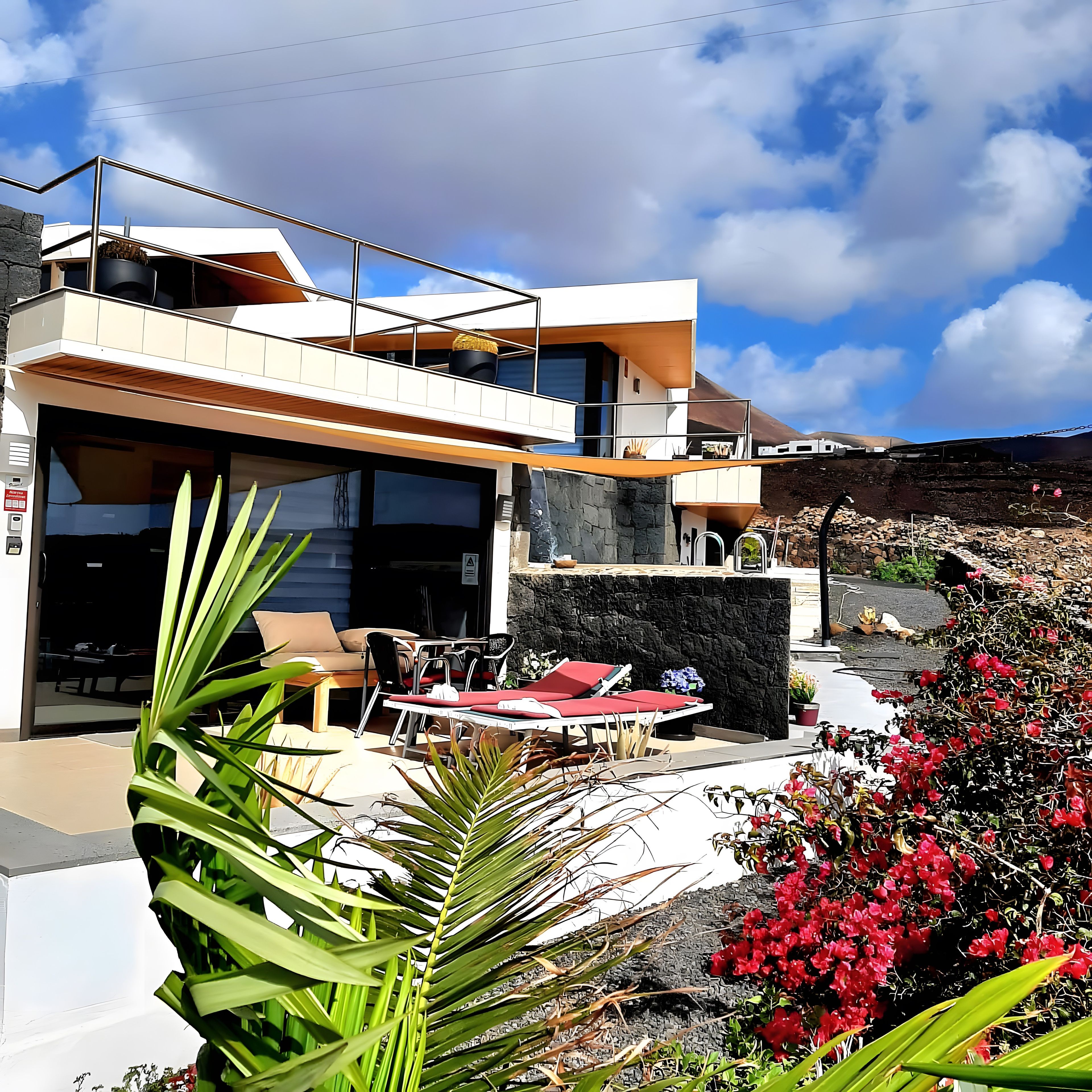 A modern two-story house with a balcony, outdoor seating, and a stone wall.