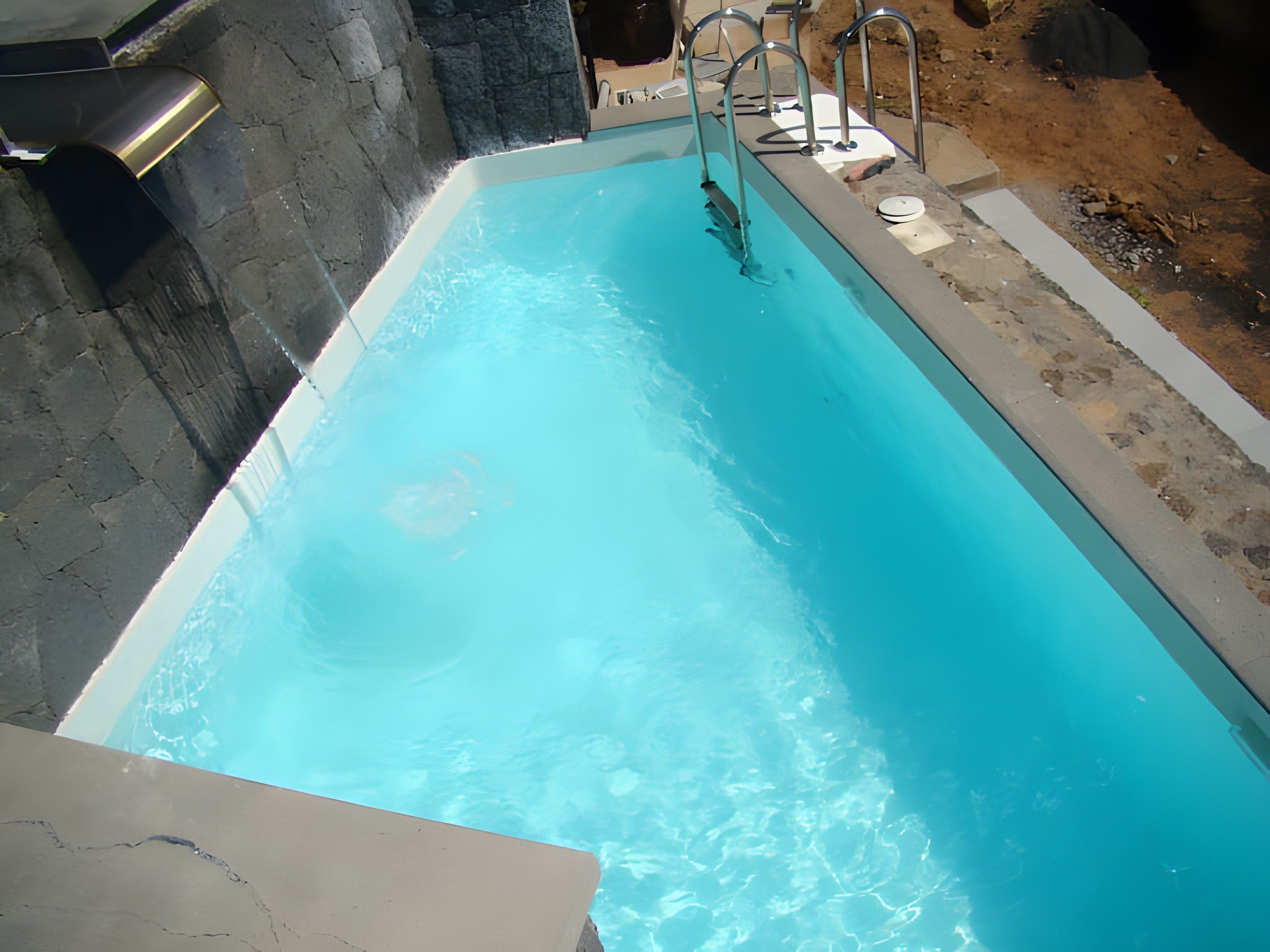 Une piscine à l’eau d’un bleu limpide, avec une échelle en métal et un mur en pierre.