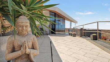 Una estatua de una figura sentada con las manos en posición de oración, un edificio moderno con grandes ventanales y una zona de patio empedrada.