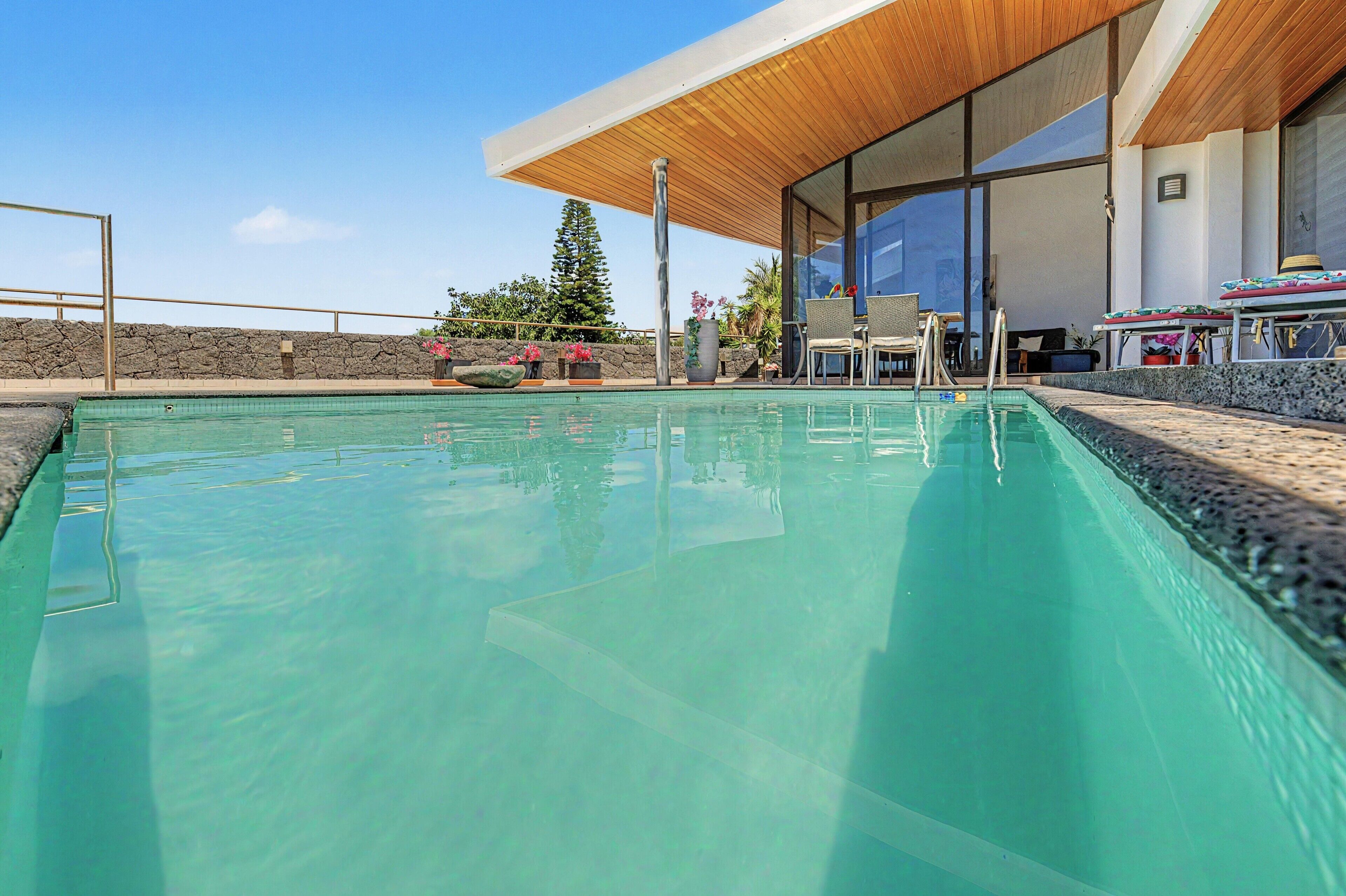 Una piscina con muelle de madera y sillas reclinables, rodeada por un muro de piedra y plantas en macetas.