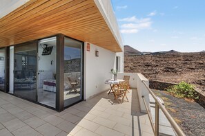 Un bâtiment moderne doté d’un plafond en bois, d’une entrée avec une porte en verre, d’une petite table avec des chaises, et offrant une vue sur un paysage aride avec des collines en arrière-plan.