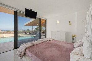 A bedroom with a large bed, a television mounted on the wall, and a view of a swimming pool and outdoor area. - Holiday Villa 'Atlantis' with Sea Views, Wi-Fi, and Air Conditioning (Las Breñas)