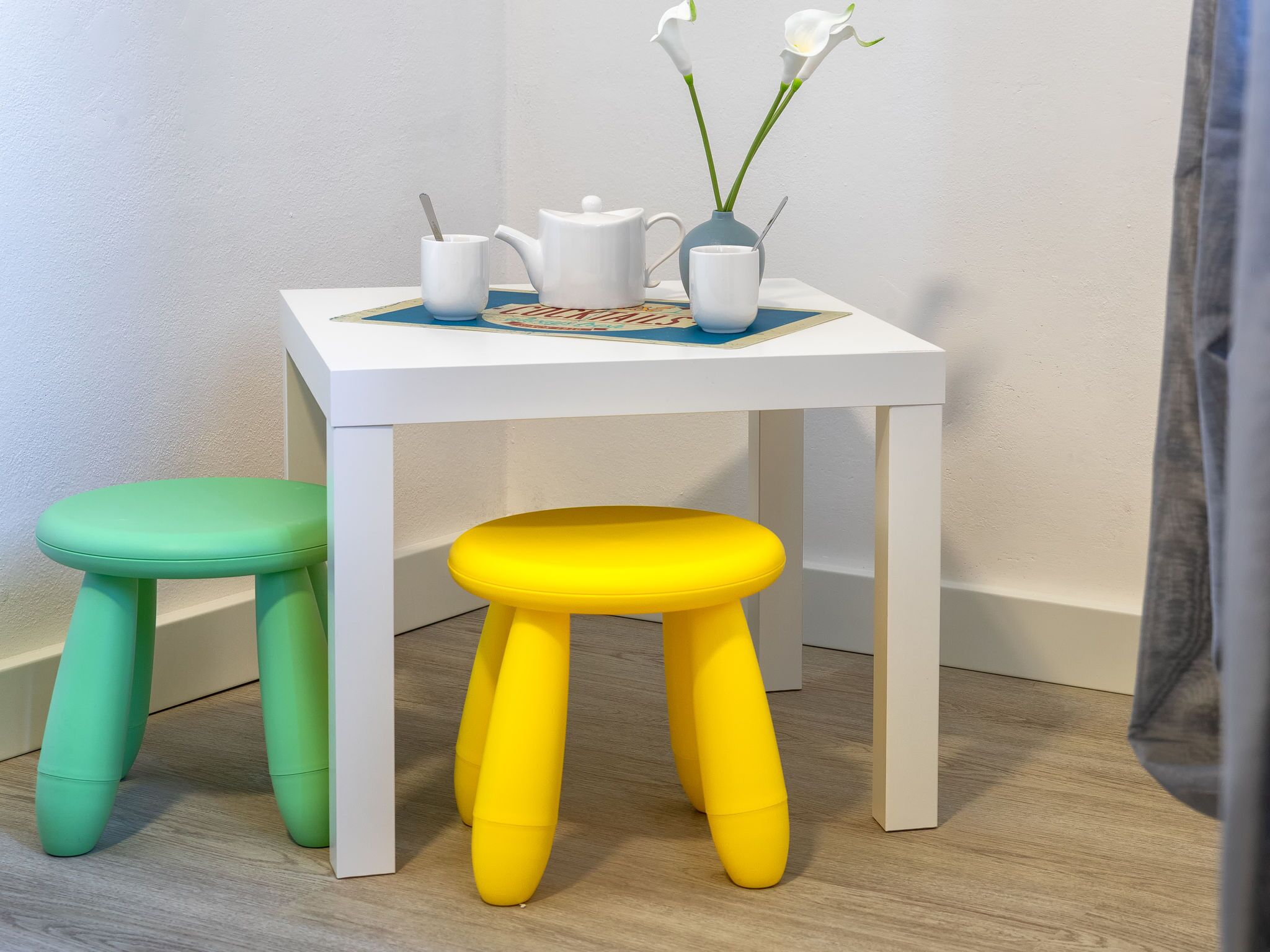 A white table with a blue placemat, a white teapot, a white mug, and a vase with white flowers. There are two stools, one green and one yellow, next to the table.