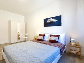 Ein Schlafzimmer mit Bett, Kleiderschrank, Stuhl, Nachttisch mit Lampe und Wandbild.