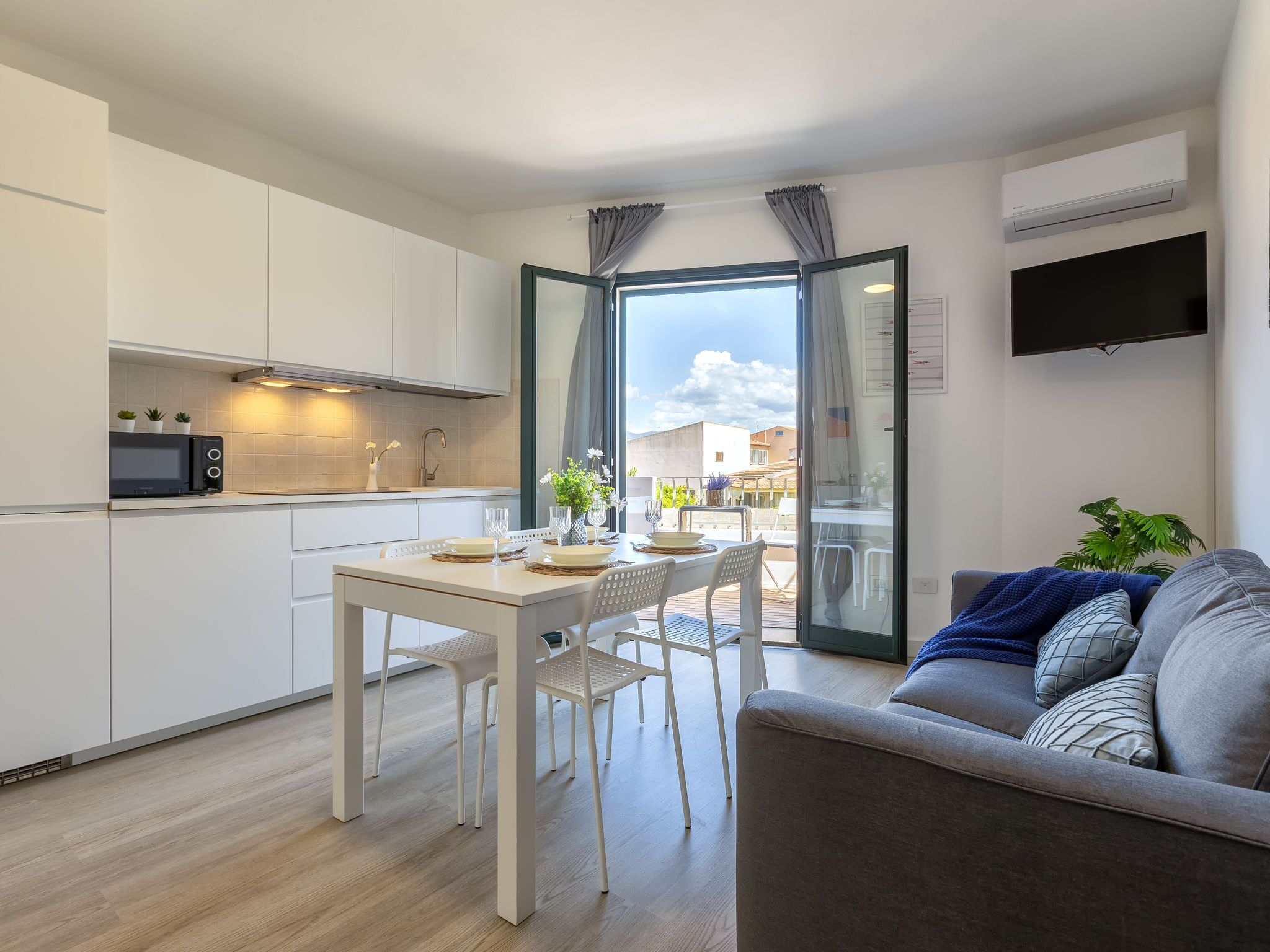 Een modern appartement met keuken, eethoek en woonkamer. Het heeft een schuifpui naar een balkon, een flatscreen-televisie aan de muur en een bank met kussens.