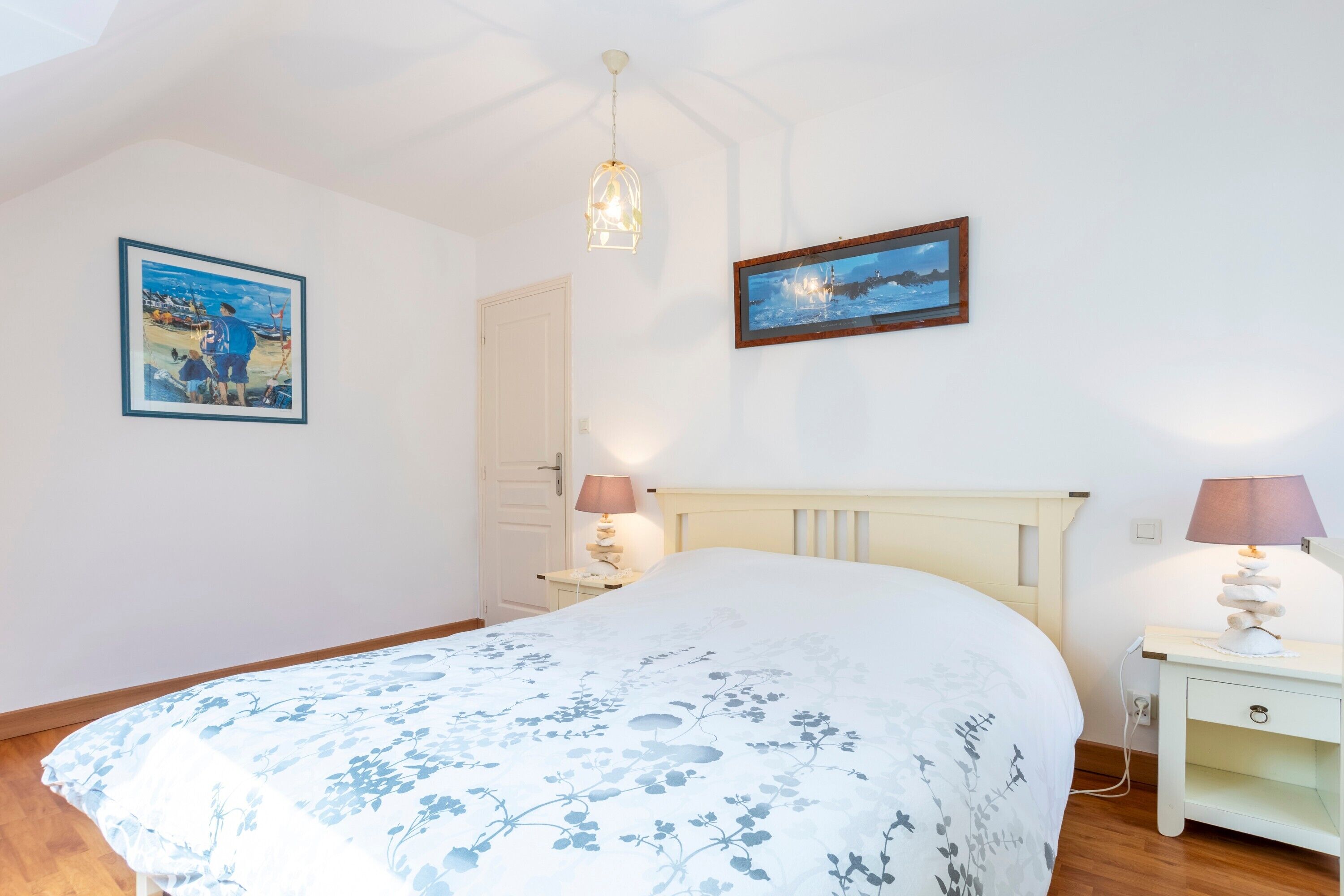 Un dormitorio con cama, dos mesitas de noche, una lámpara colgante y dos cuadros en la pared.