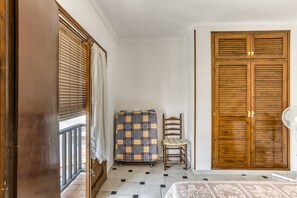Une pièce comprenant un lit, une armoire en bois, une chaise, un objet recouvert d’un tissu et une fenêtre avec des stores.