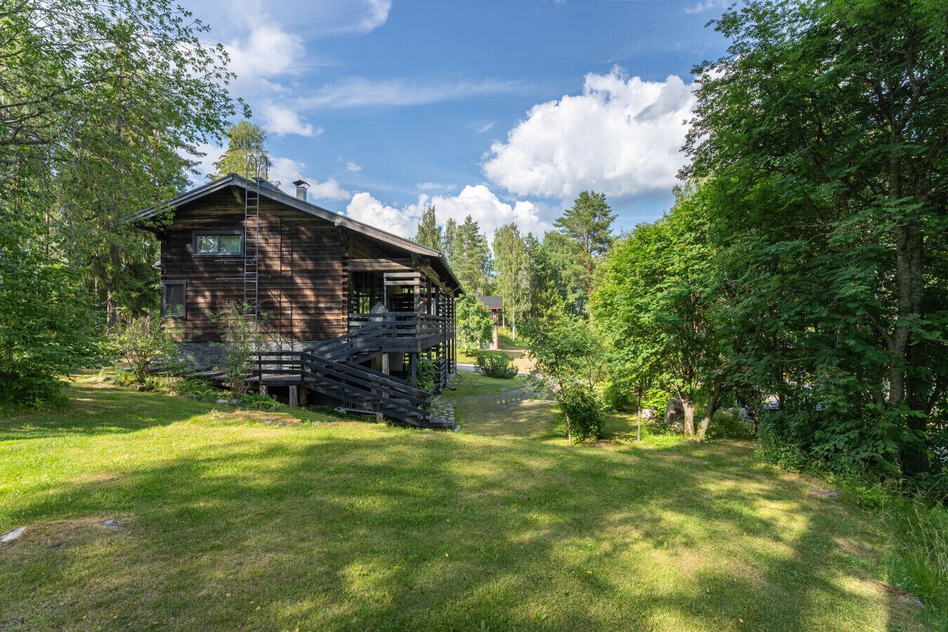 Een rustieke houten cottage met een houten trap naar een veranda, omgeven door weelderig groen en bomen.