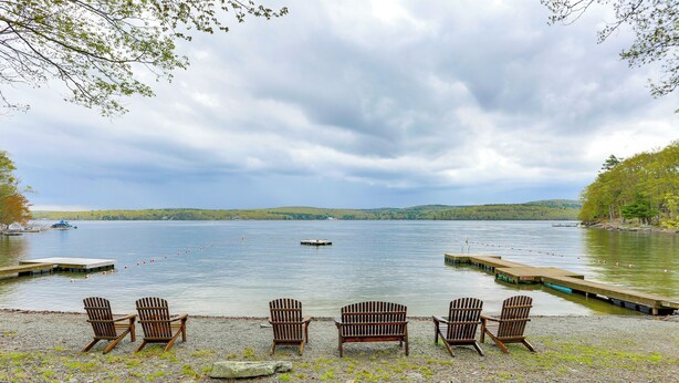 Blick auf einen See mit Holzstühlen, die zum Wasser zeigen, einem Steg und einem Boot in der Ferne.