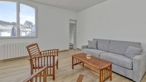 A modern living room with a grey sofa, wooden chairs, a wooden table, and a large window.