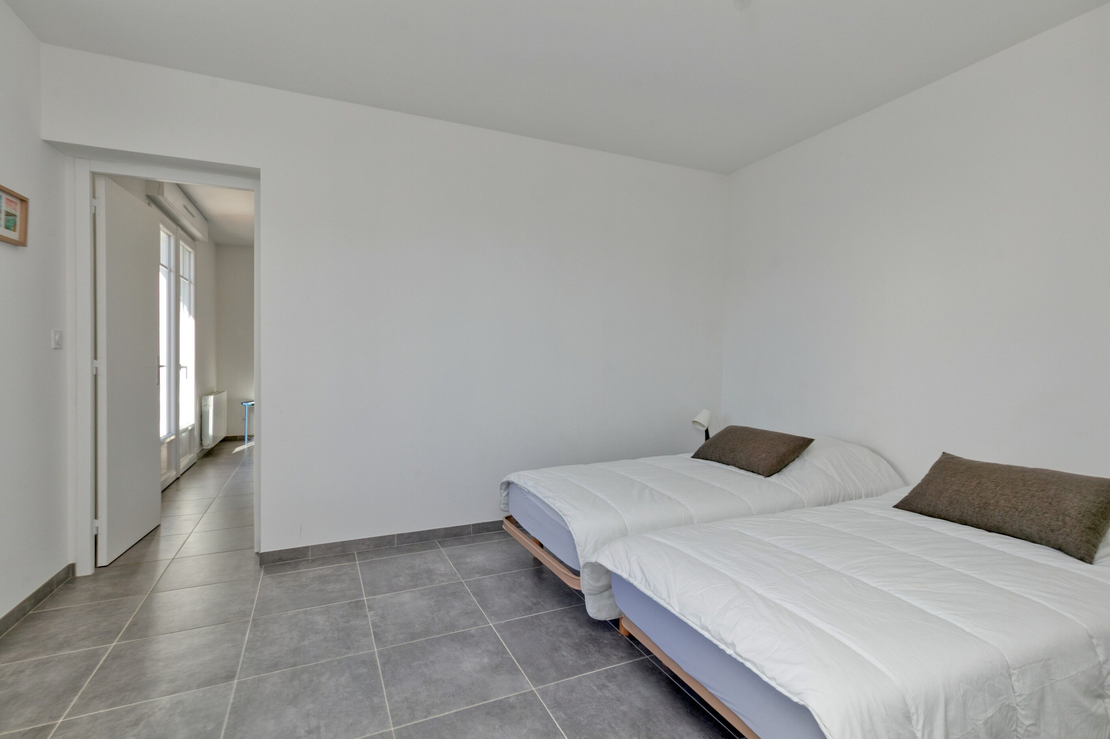 A bedroom with two beds, grey tiled flooring, and a hallway leading to another room.