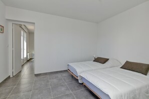Une chambre avec deux lits, un sol carrelé de couleur grise et un couloir menant à une autre pièce.