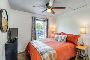 Une chambre à coucher avec un lit, un ventilateur de plafond, un téléviseur posé sur un meuble, une lampe et une fenêtre avec des rideaux.