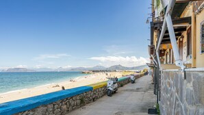 A coastal street with a stone wall, a scooter parked on the sidewalk, and a beach in the background.