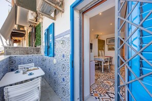 A narrow outdoor corridor with a blue door, a white table with chairs, and patterned tiles on the walls.
