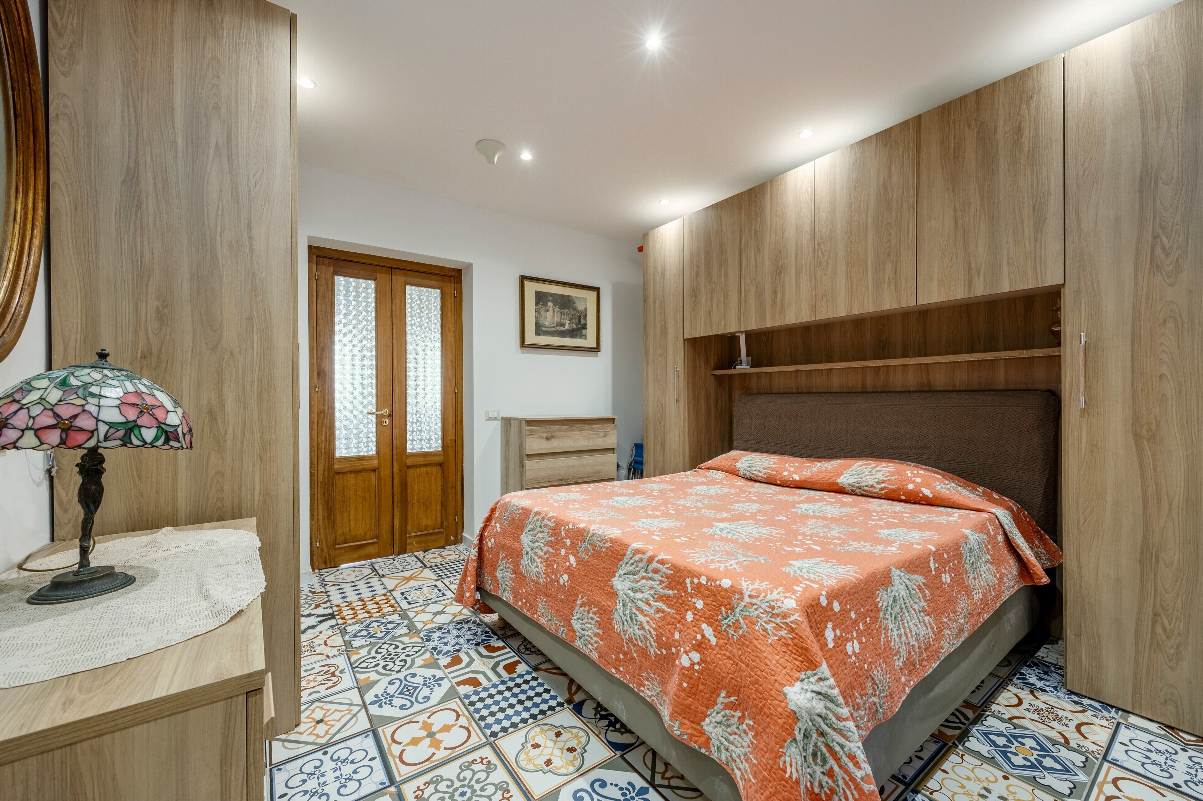 A bedroom with a bed, a nightstand, a lamp, a door, and a wall-mounted shelf.