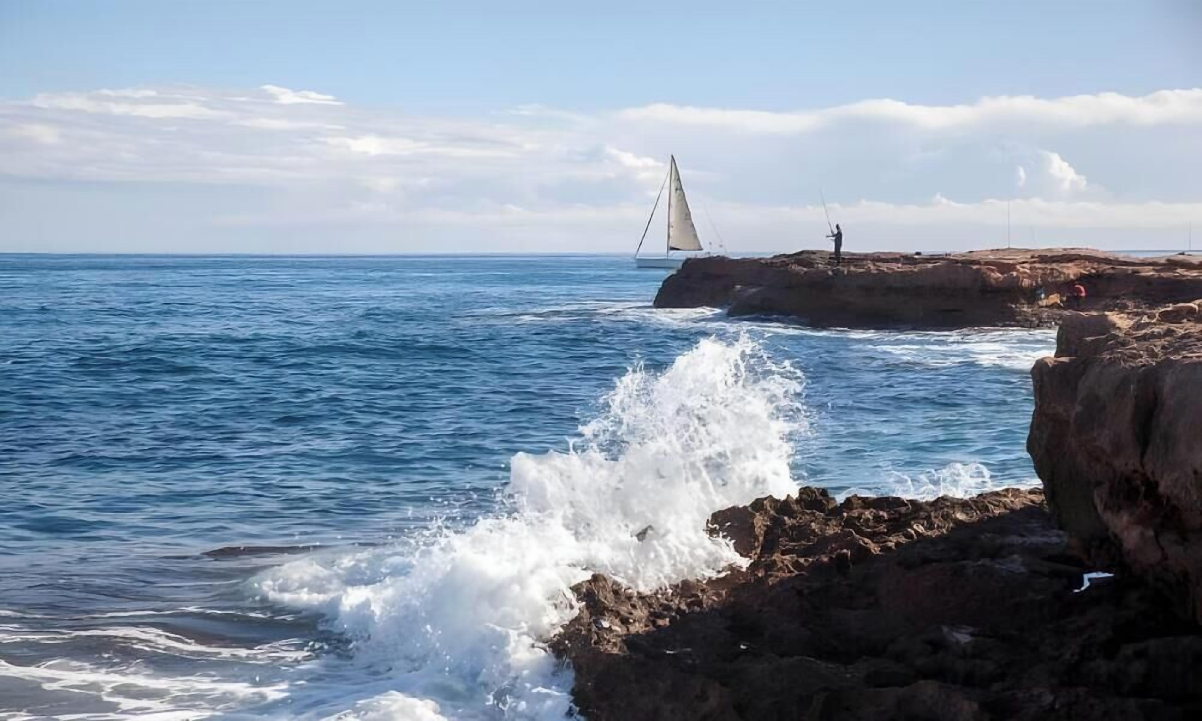 Escena costera con olas rompiendo contra rocas, un velero a lo lejos y una persona de pie sobre las rocas.