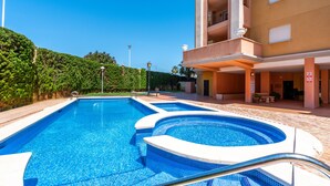 Una piscina con borde de azulejos, un edificio con balcones y un cielo azul despejado.