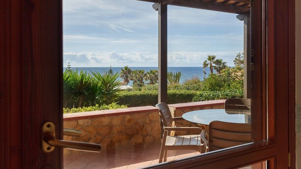 Vue depuis une pièce dont le mur est en pierre et la porte en bois, donnant sur une terrasse avec une table et des chaises, des palmiers et l’océan.