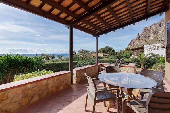A covered outdoor area with a table and chairs, stone walls, and a view of the sea and mountains.