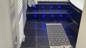 A modern bathroom with a bathtub, a white sink, and a shower. There is a towel rack with towels, a patterned rug on the floor, and blue LED lights under the bathtub. - QUIET HOUSE (Cuxac-d'Aude)
