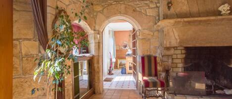A stone-walled hallway leading to a room with a fireplace, a tiled floor, and a plant.