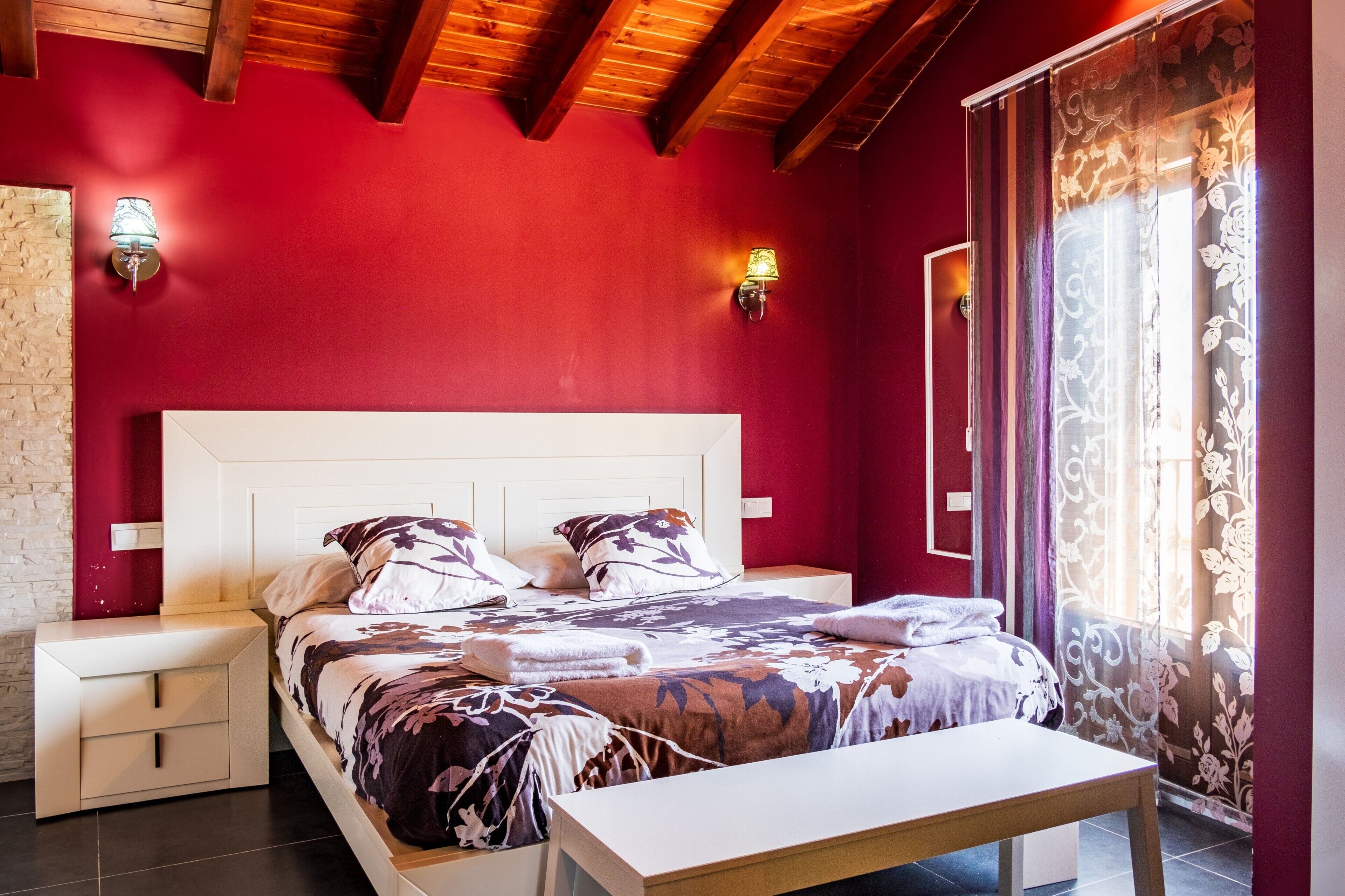 Ein Schlafzimmer mit roter Wand, einem weißen Bett, zwei Nachttischen und einem Fenster mit Vorhängen.