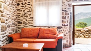 Une pièce aux murs de pierre, avec un canapé, une table et une vue sur un mur de pierre et la verdure à l’extérieur.