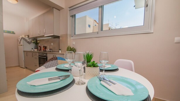 A dining area with a round table set for two, a kitchen in the background, and a window with a view of buildings. - Apartment 'Mantisses Studio' with Shared Terrace, Wi-Fi and Air Conditioning (?a?aµ?ta)